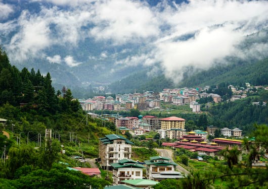 Colorful houses with green and red roofs sprawl along the Thimphu Valley in Bhutan. The Valley is lined with green trees and hills, and a cloudy fog is coming over the hills onto the city.