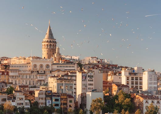 The Galeta tower is shown at sunset in Istanbul, Turkey, with a flock of birds flying in the sky.