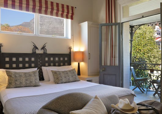 A hotel room in South Africa is shown with a comfortable double bed and French doors opening onto a patio with two chairs. There is a green tree out near the patio and hills out the windows above the bed.