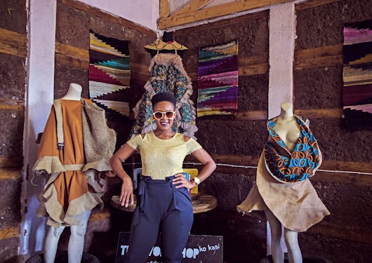A Black woman stands in a local curio textile shop, wearing yellow sunglasses and smiling at the camera. Patterned textiles are on mannequins next to her and hanging up behind her. This is in South Africa.