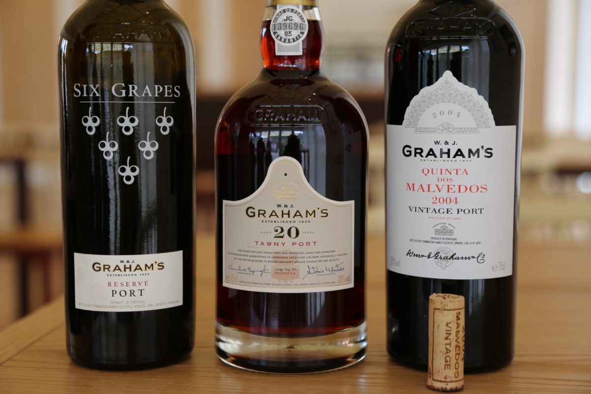 Three dark wine bottles sit on a wooden table in a horizontal line, with a beige cork standing upright in front of the bottle on the right. The bottles are Port wines from Portugal.