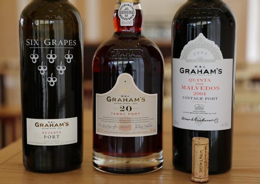 Three dark wine bottles sit on a wooden table in a horizontal line, with a beige cork standing upright in front of the bottle on the right. The bottles are Port wines from Portugal.