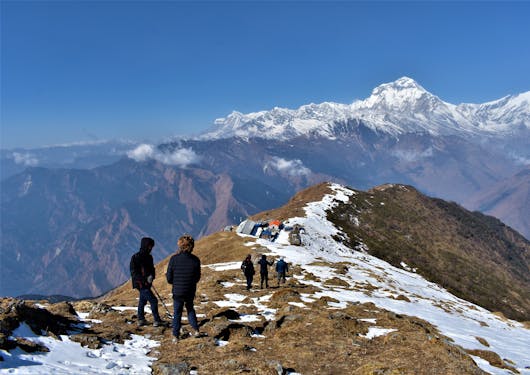 Atop a ridge, there are a couple of people in the foreground, and three more in the background. The ground is brown with a path of snow. Snow-capped mountains are in the distance. It's a blue sky, sunny day.