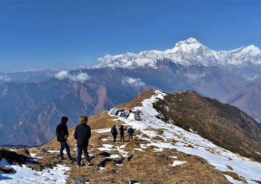 Atop a ridge, there are a couple of people in the foreground, and three more in the background. The ground is brown with a path of snow. Snow-capped mountains are in the distance. It's a blue sky, sunny day.