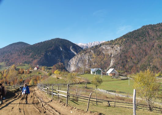 A woman walks along a dirt road in Romania's Carpathian Mountains on a sunny day. Tree-covered hills and some snow capped mountains are in the background.