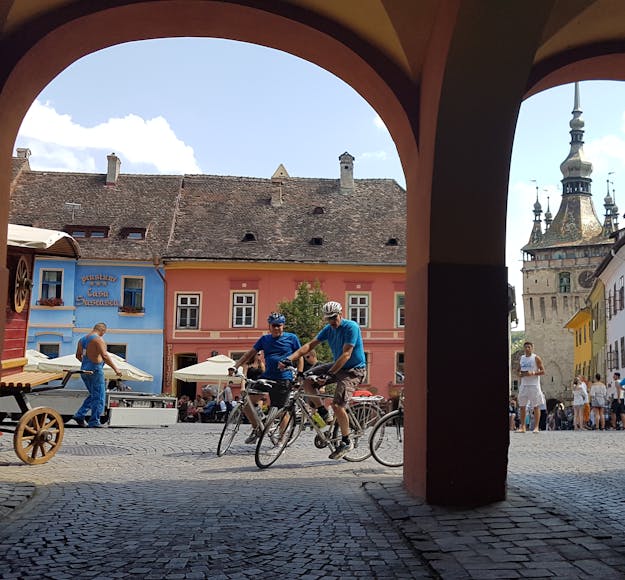 A couple of adult Caucasian bikers pull into a colorful town in Romania; there are arches in the shade in the foreground and colorful buildings lining the square in the background, with various people walking and standing around.
