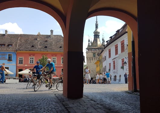 A couple of adult Caucasian bikers pull into a colorful town in Romania; there are arches in the shade in the foreground and colorful buildings lining the square in the background, with various people walking and standing around.