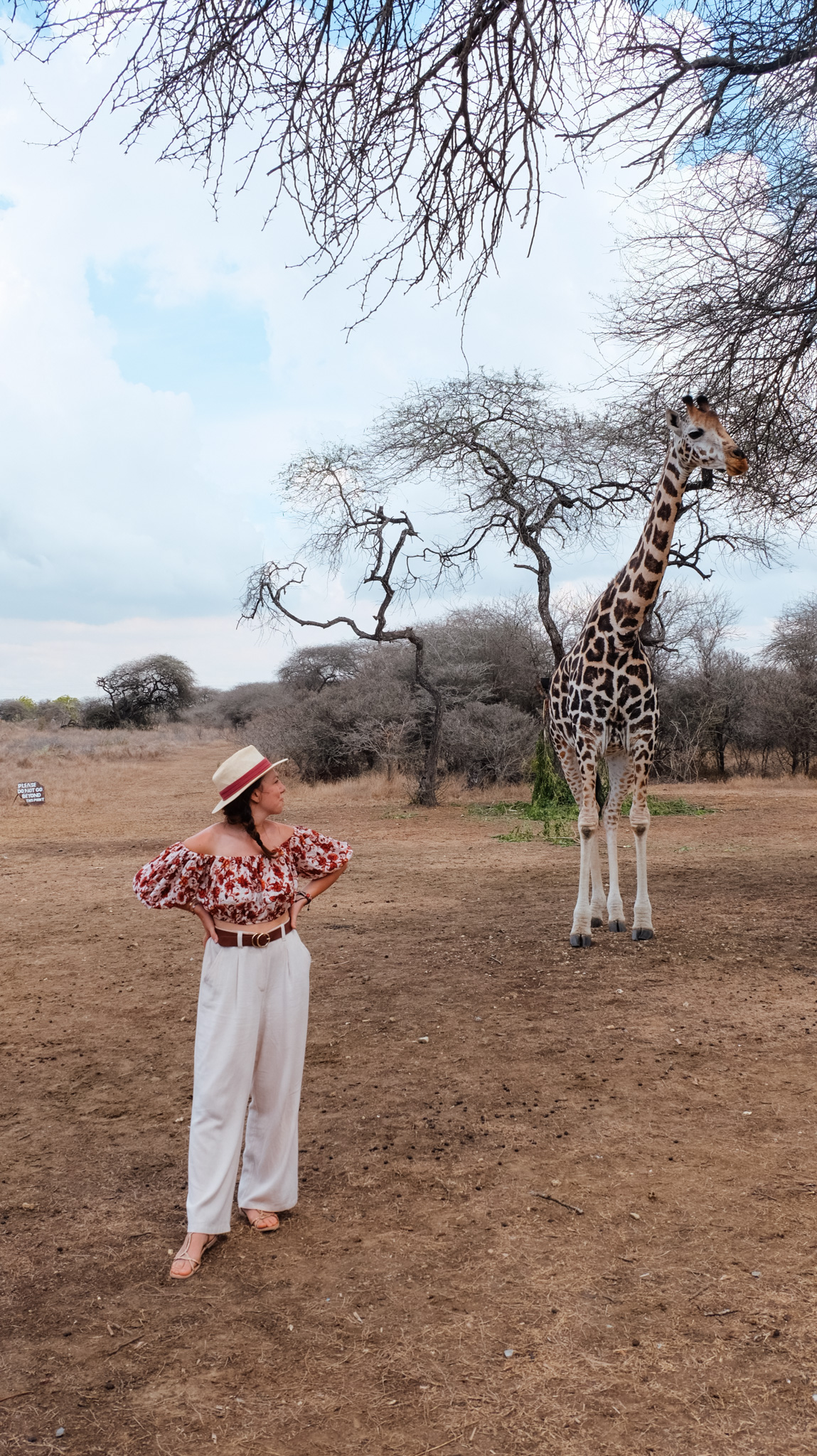A young woman stands and looks over her shoulder at a towering nearby giraffe.