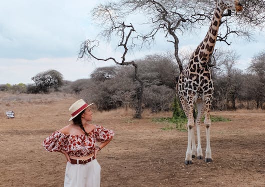 A young woman stands and looks over her shoulder at a towering nearby giraffe.