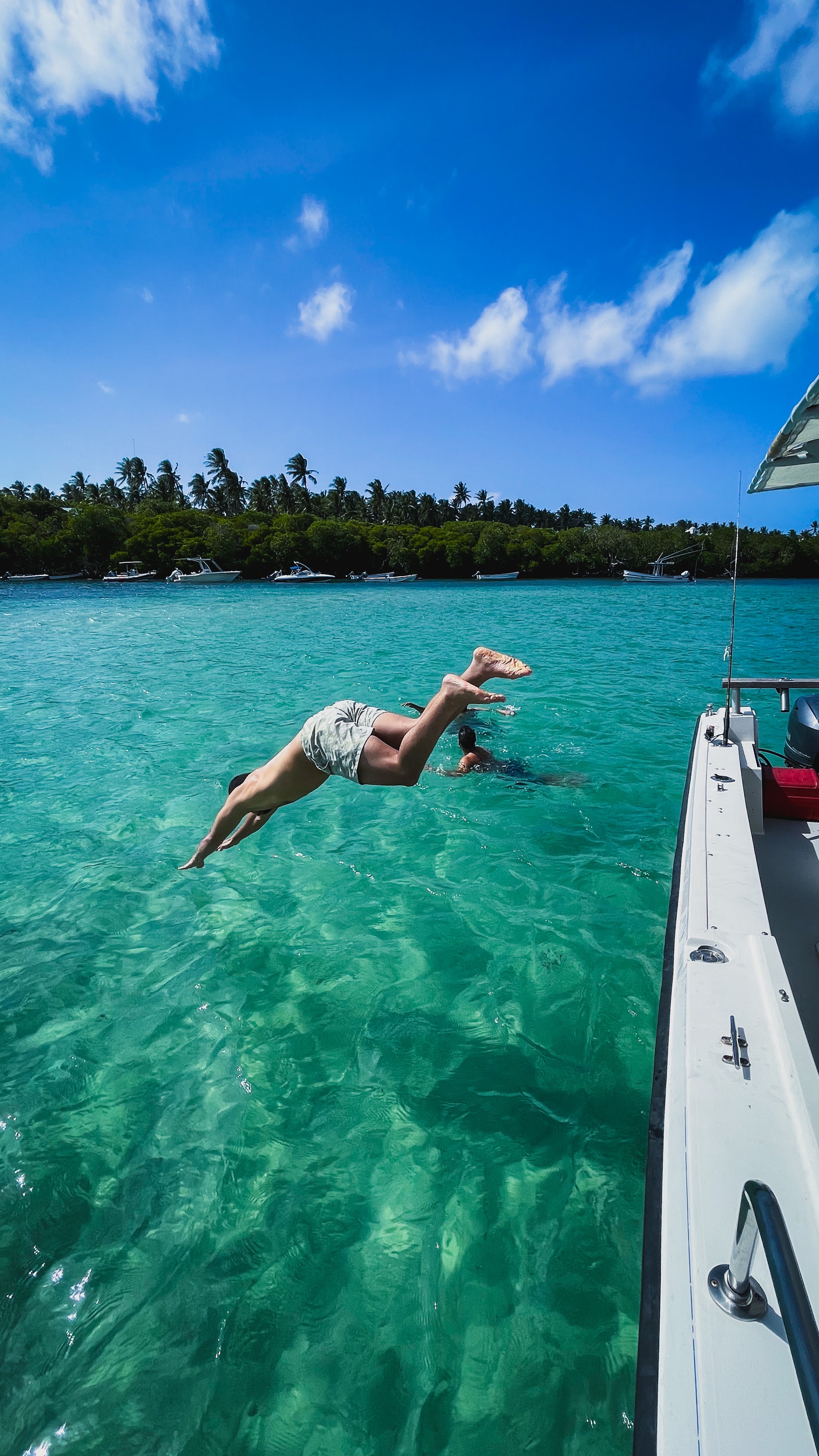 A young white man jumps off the side of a boat into bright teal water on the Kenyan Coast, with green trees in the background and underneath a blue sky.