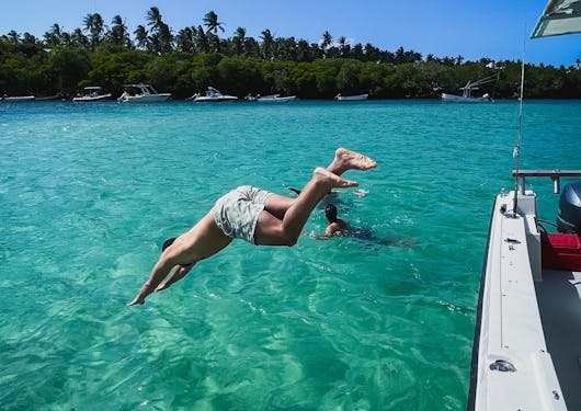 A young white man jumps off the side of a boat into bright teal water on the Kenyan Coast, with green trees in the background and underneath a blue sky.
