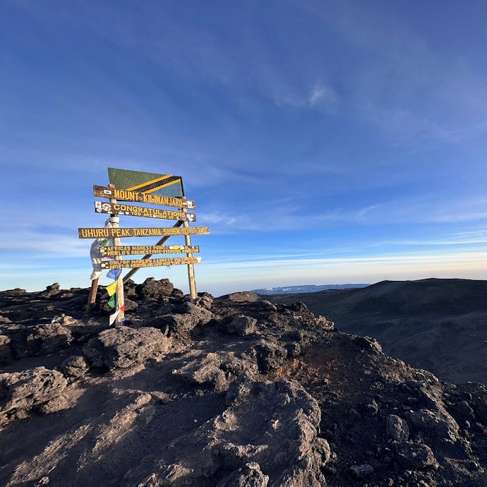 At the summit of Mount Kilimanjaro in Tanzania, a wooden sign post stands indicating Uhuru Peak, the highest point in Africa! The sign post has yellow lettering and a Tanzanian flag above it, with some prayer flags on the left hand side. The sign is in the left part of the frame, with the view over the mountain tops to the right. Blue sky and sunny, with some wispy clouds.
