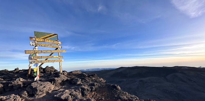 At the summit of Mount Kilimanjaro in Tanzania, a wooden sign post stands indicating Uhuru Peak, the highest point in Africa! The sign post has yellow lettering and a Tanzanian flag above it, with some prayer flags on the left hand side. The sign is in the left part of the frame, with the view over the mountain tops to the right. Blue sky and sunny, with some wispy clouds.