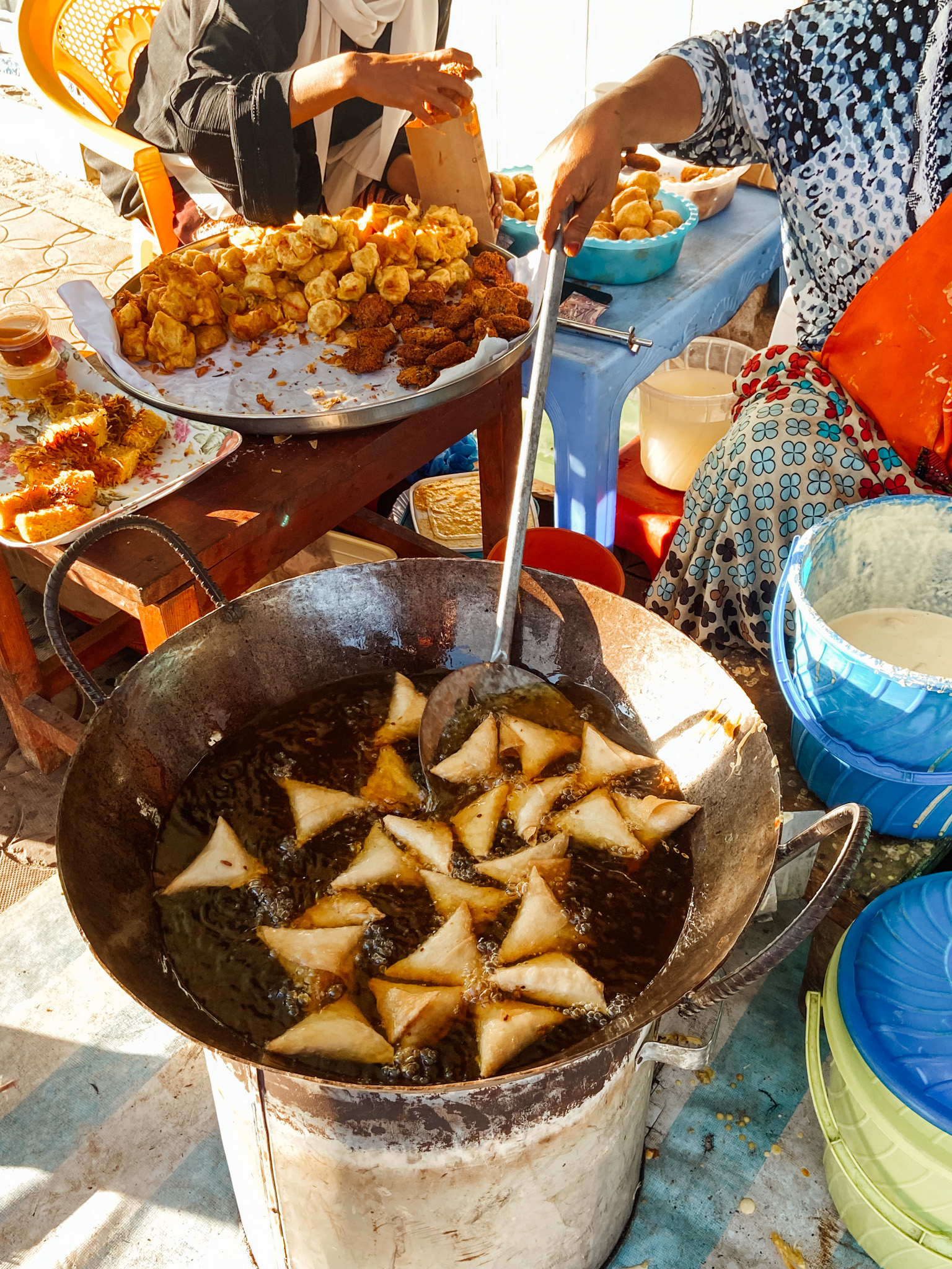 A local Kenyan woman standing out of frame cooks triangular-shaped samosas in a big skillet of oil, as another person is also out of frame preparing other local food.