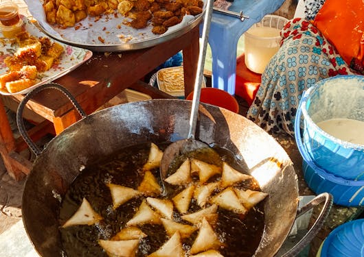 A local Kenyan woman standing out of frame cooks triangular-shaped samosas in a big skillet of oil, as another person is also out of frame preparing other local food.