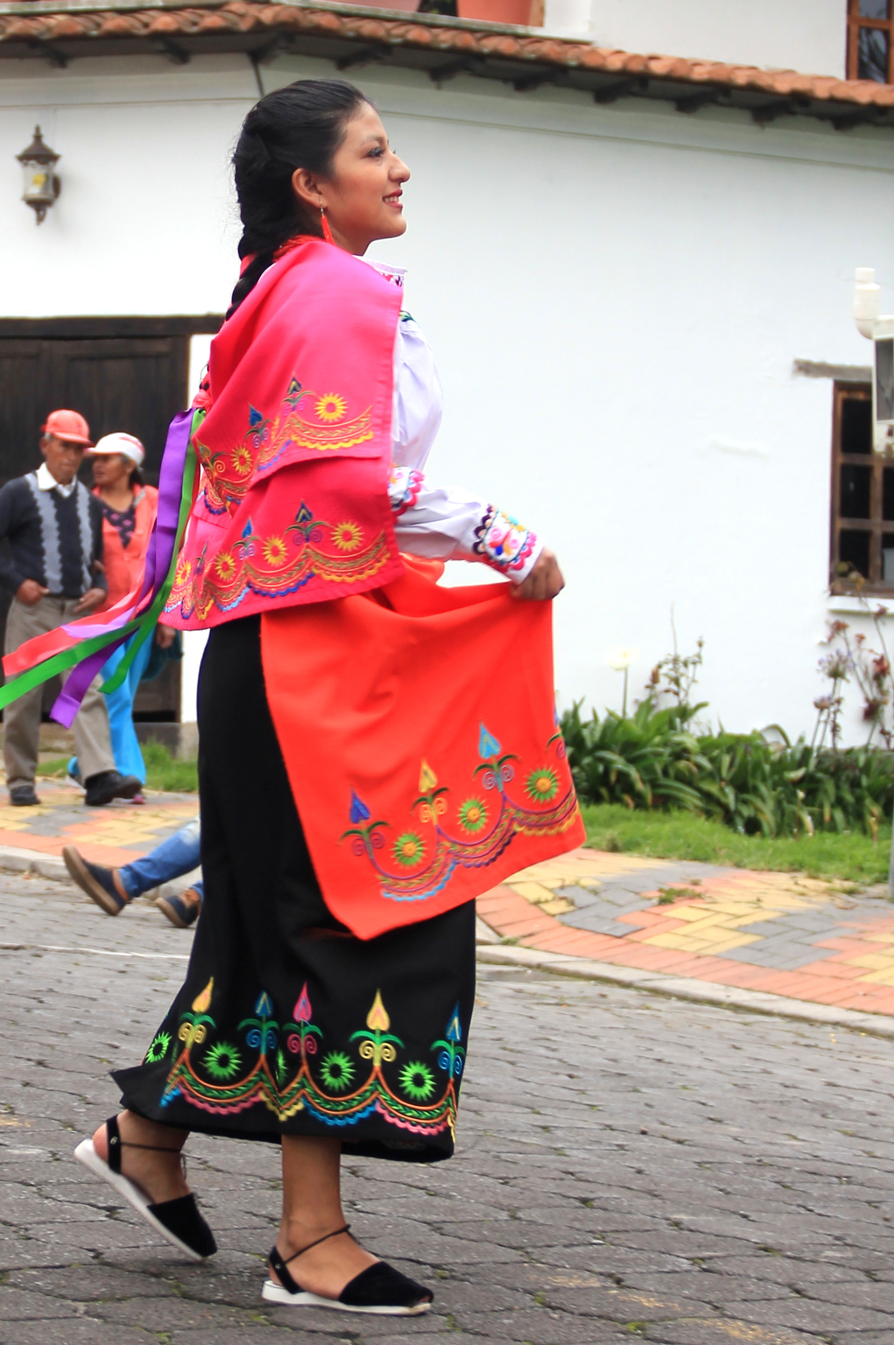 An indigienous Ecuadorian woman stands tall, in black shoes, a black skirt with colorful embroidery along the bottom, a long sleeve white shirt and a bright red shawl embroidered along the bottom, matching the skirt. Her dark hair is braided and she is walking and smiling.