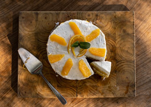 A typical South African dessert, an orange cake, is shown from above; it rests on a wooden platter on a wooden counter with a silver serving spoon off to the left. The circular cake has white frosting and is adorned with slices of fresh oranges. One piece is slightly cut and removed.