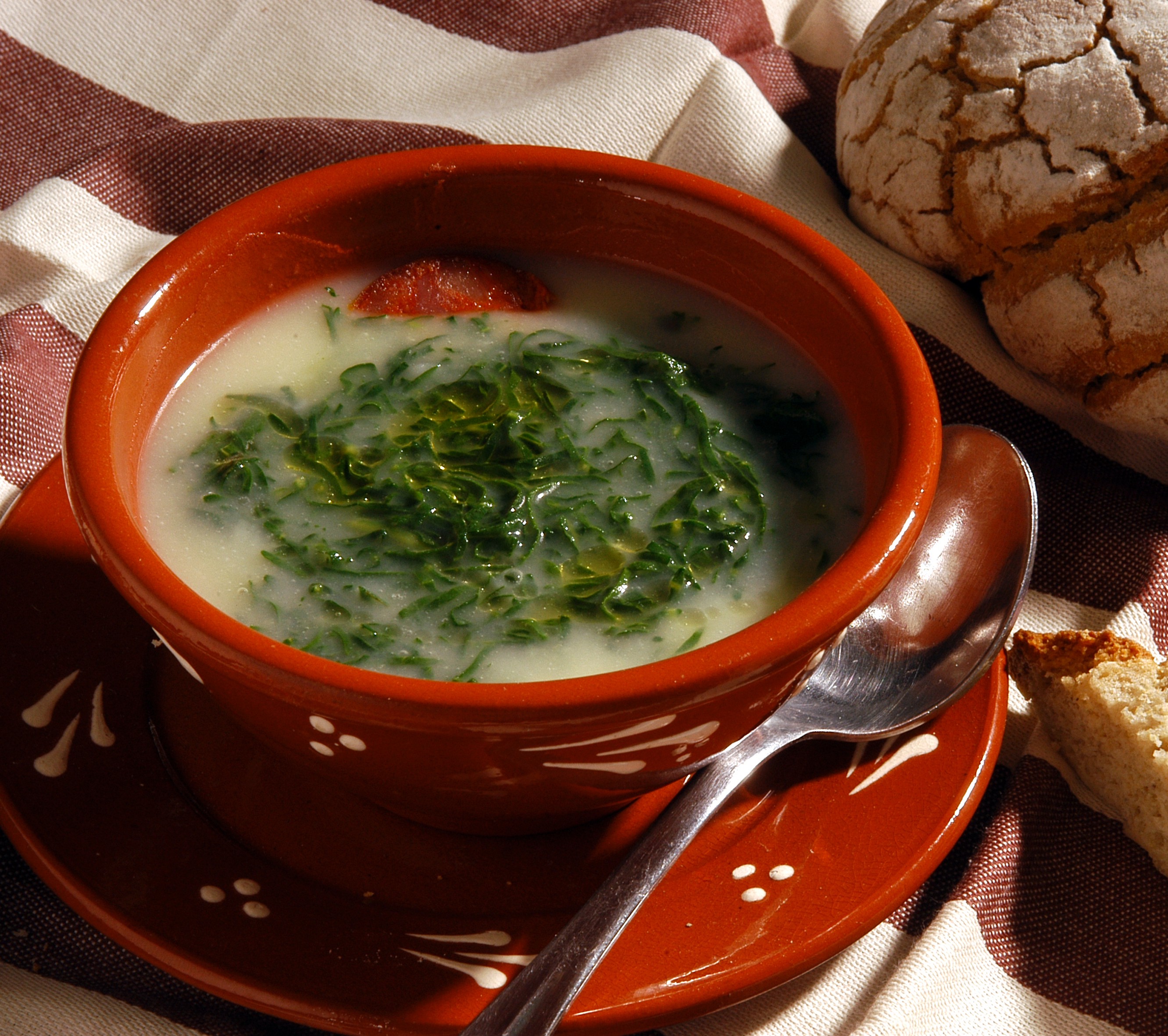 A fresh bowl of Caldo Verde soup rests in a traditional, red-painted Portuguese bowl. A load of bread sits nearby.