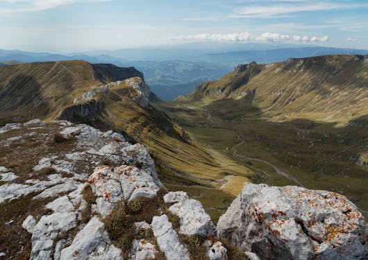 The Bucegi Mountains in Romania are shown in partial shade and partial sun, with rocky plateaus on the left and right, and rolling hills in the background. Big boulders covered in orange moss are in the foreground. A path is weaving between the two plateaus.