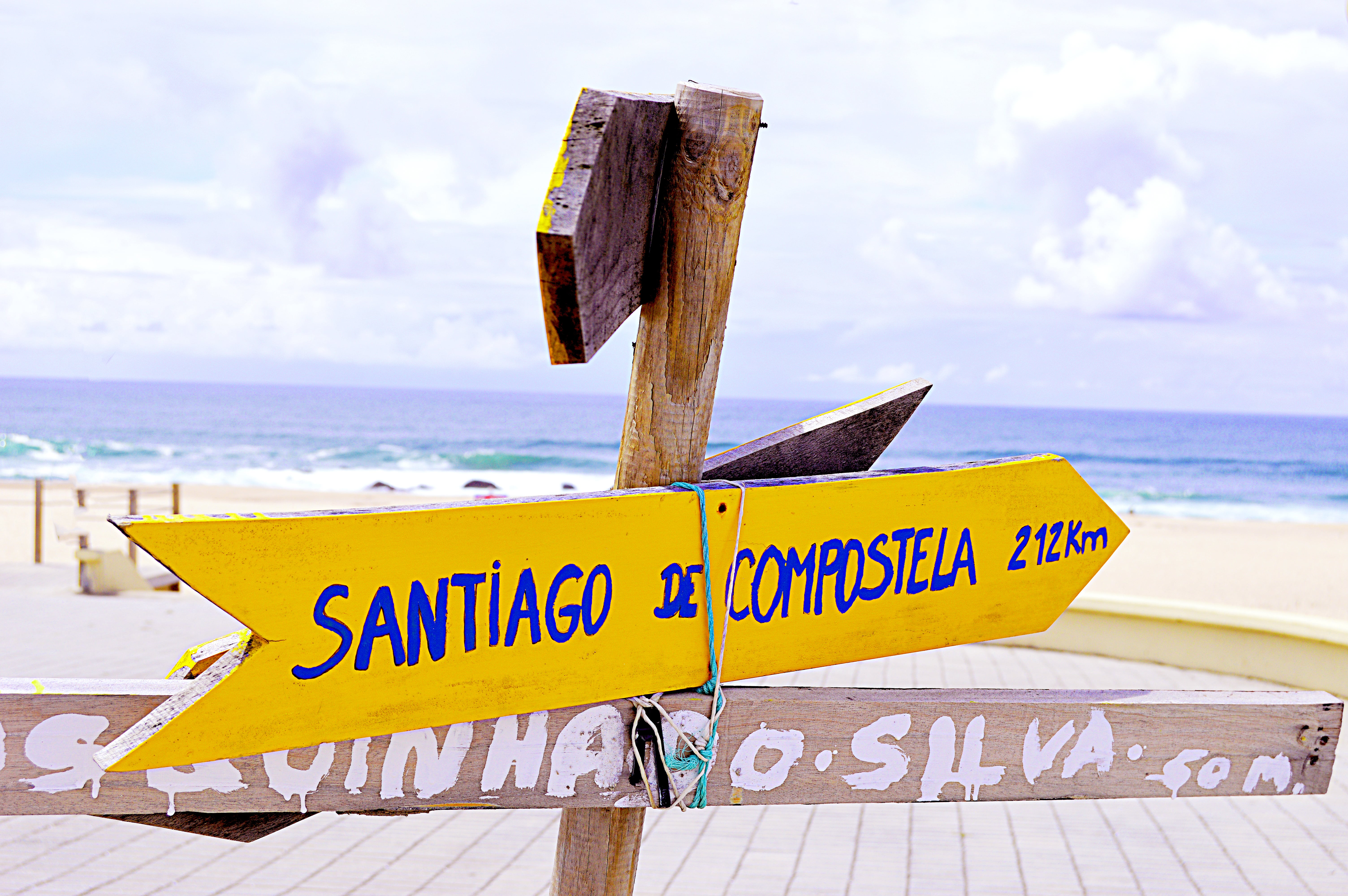 A yellow signpost with blue lettering stating "Santiago de Compostela: 212 km" is tied to a wooden signpost. The Atlantic Ocean is in the background just after a white sand beach.