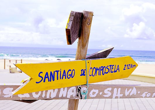 A yellow signpost with blue lettering stating "Santiago de Compostela: 212 km" is tied to a wooden signpost. The Atlantic Ocean is in the background just after a white sand beach.
