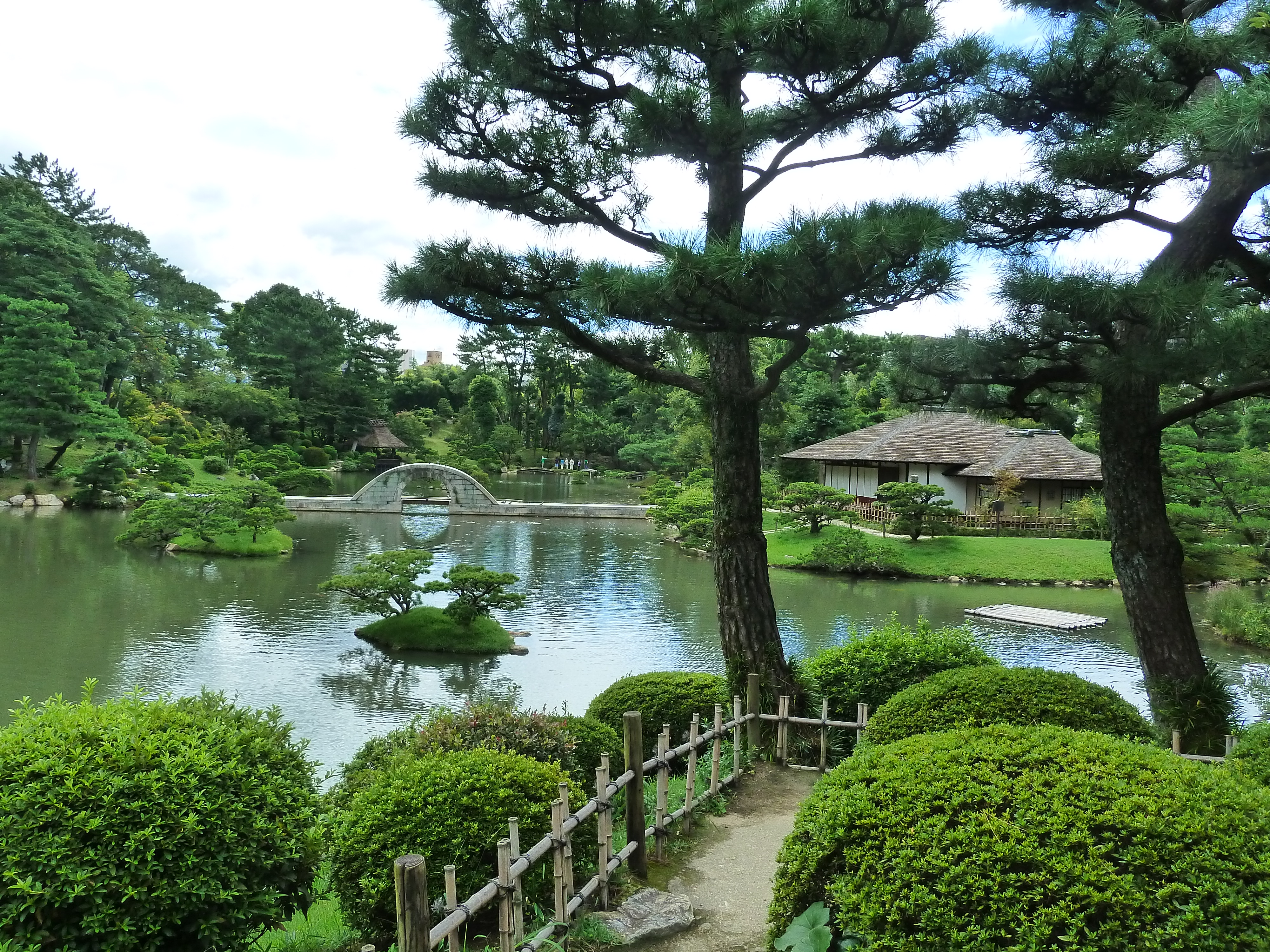 A zen garden in Japan is shown with greenery surrounding a pond, a bridge in the foreground, and an overcast sky.
