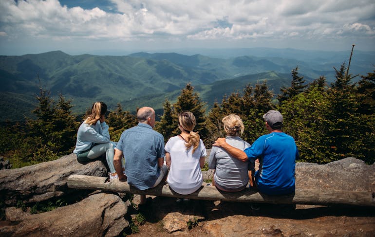 A Caucasion woman sits on a rock, next to a group of 4 Caucasian people sitting on a log; they are all looking out over a landscape of pointy green hills and a cloudy yet bright sky. Green trees are in the foreground just past the lookout point.