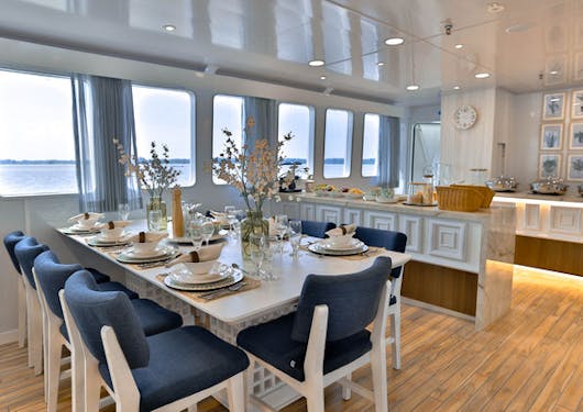 The dining area with a table and chairs in front of windows is shown on the luxury yacht used for the Galapagos Citizen Science scuba diving trip.