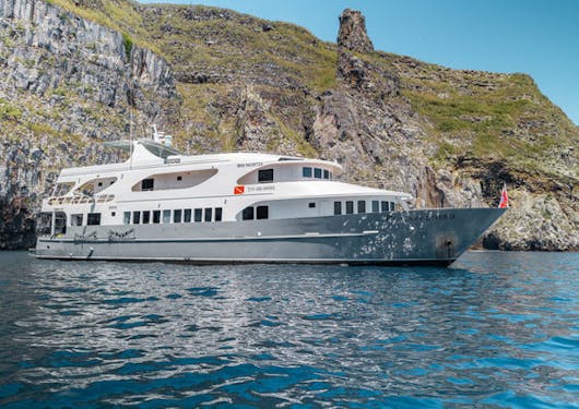 A white luxury yacht resting in front of a large, grass covered hill is shown on the luxury yacht used for the Galapagos Citizen Science scuba diving trip.