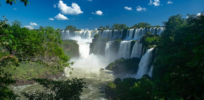 Argentine side of Iguazú Falls (Parque Nacional Iguazú) from the lower trail - a beautiful waterfall overflowing with white ribbons with greenery in the front left of the frame and a pure blue sky with a few white clouds above.
