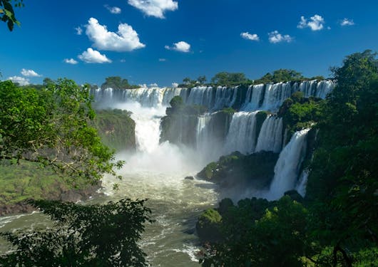Argentine side of Iguazú Falls (Parque Nacional Iguazú) from the lower trail - a beautiful waterfall overflowing with white ribbons with greenery in the front left of the frame and a pure blue sky with a few white clouds above.