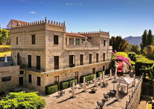 A three story luxury hotel in Portugal stands proudly in the sun with an open terrace, greenery around, and a blue sky.