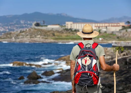 A walker along the Portuguese Coastal Way Camino rests with his walking stick in his right hand, a red backpack, and a tan hat, standing and looking over a rugged beach in northern Portugal.