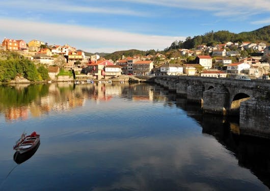 A low-lying bridge traverses a calm body of water with a village on the other side in the sunshine.