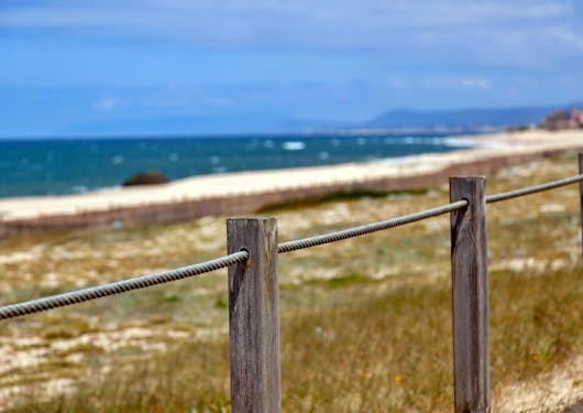 A wood and wire railing is in focus on a sunny day with the Portuguese Coast and Atlantic Ocean out of focus in the background.