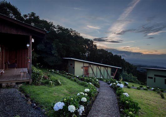 An intimate lodge is nestled in the coffee lands of Costa Rica, with a lined green path leading up to it and a sunset in the background.