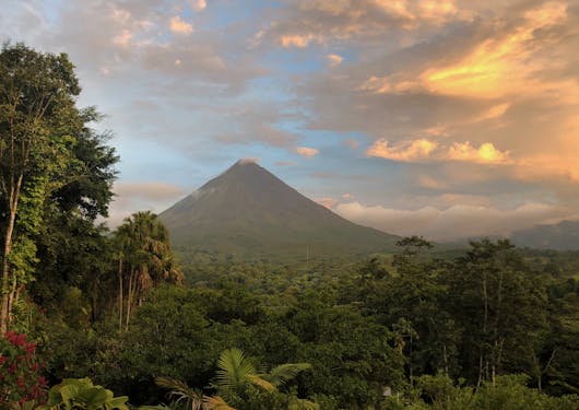 The Arenal Volcano of costa Rica is shown towering over trees as the sun sets to the right.