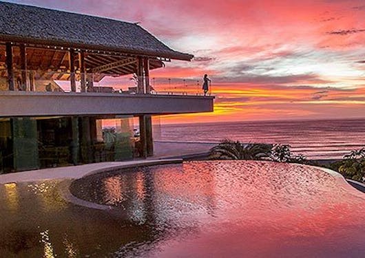 A lodge is shown in Nosara, Costa Rica against a pink and red-hued sunset.
