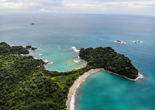 An aerial photo of Manuel Antonio is shown from above, with blue and teal water hugging the forested Pacific Ocean coastline in Costa Rica.