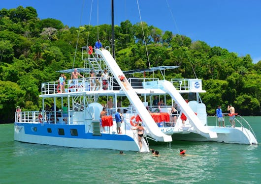The Catamaran tour in Manuel Antonio in Costa Rica is shown on green water in front of bright green trees on land and a blue sky. It's a double decker and a guest is sliding off of the top deck into the water.