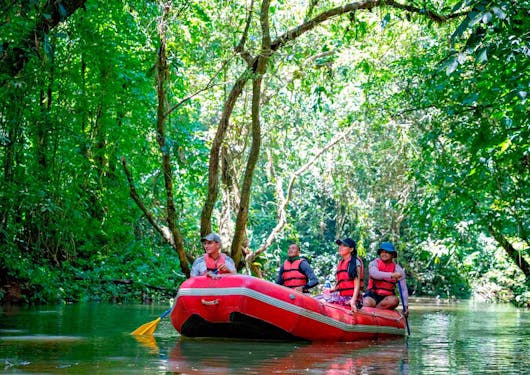 Several travelers wearing red life jackets are on a red inflatable raft floating down a river in Costa Rica, surrounded by lush greenery.