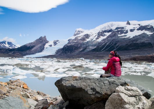 A woman in warm clothing gear sits on top of a rock overlooking an ice-filled lagoon with an Argentine glacier in the background.