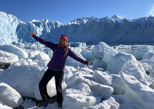 An adult in warm gear clothing stands upon a white ice field in Argentina with their arms extended in glee, under a bright blue sky.