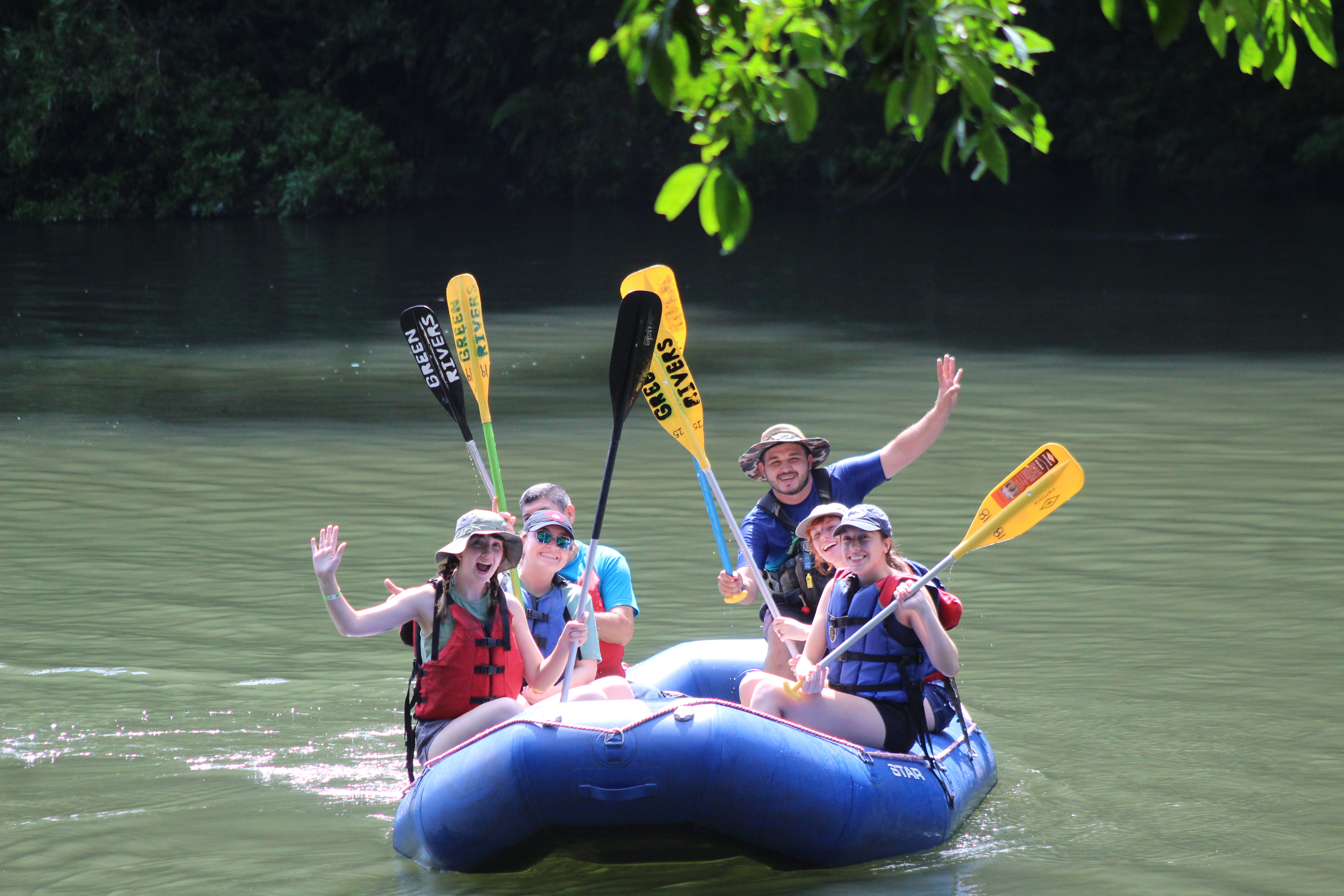 A group of 6 adults in life jackets sit atop a sturdy, inflatable raft as they drift down the river in Costa Rica, gleefully raising their yellow-tipped paddles in the air.