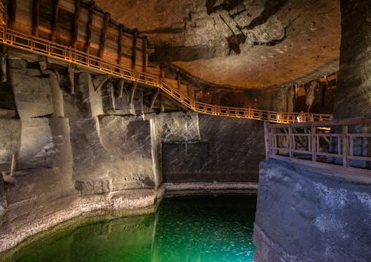 The inside of the Wieliczka salt mines near Krakow, Poland are shown in a dimly lit setting, with a pool green water on the ground.