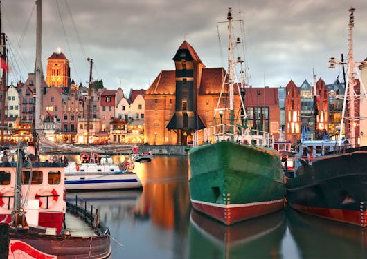 The port of Gdansk, Poland is shown with several colorful boats in the foreground at dusk with the warmly lit city in the background.