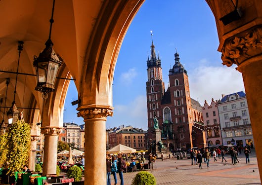 The main square of Krakow, Poland is shown from underneath some orange archways with a church in the distance set against a blue sky.