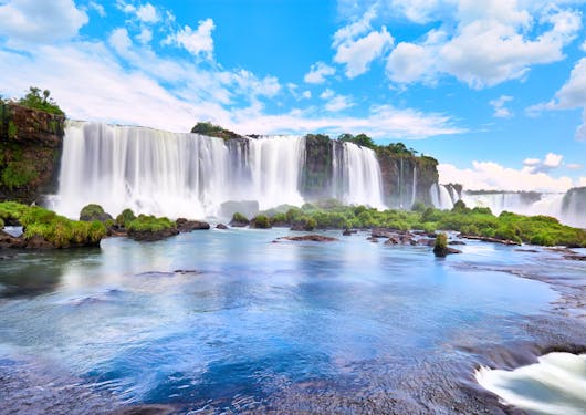 The stunning Argentine Falls in Iguazu are shown with their dazzling cascades under a blue sky with white clouds.