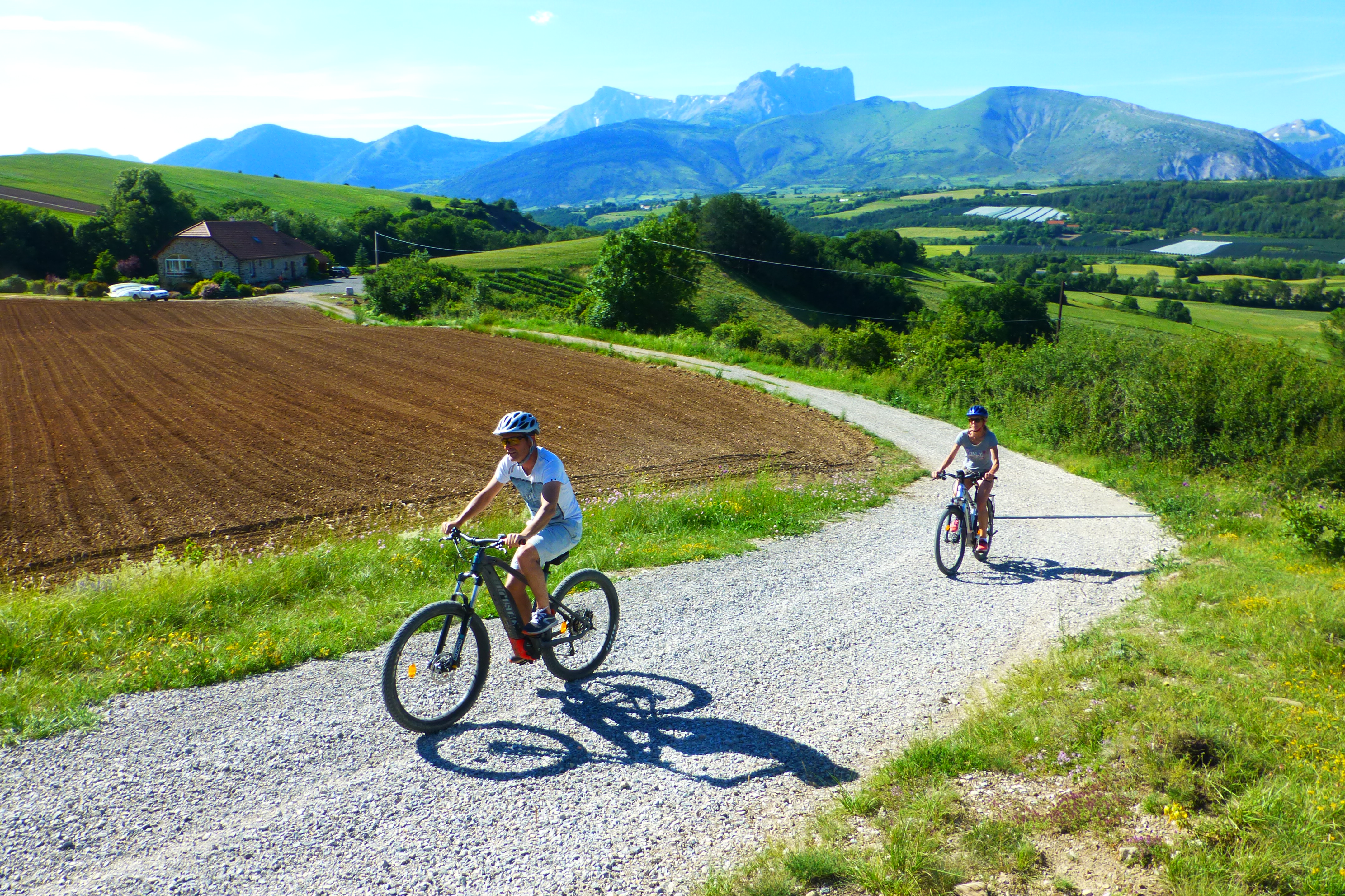 Two bikers cycle along a gravel path in the French Alps on a sunny day, with a brown field to the left and mountains in the background. This is in the French Alps on a sunny day on a sustainable cycling tour.