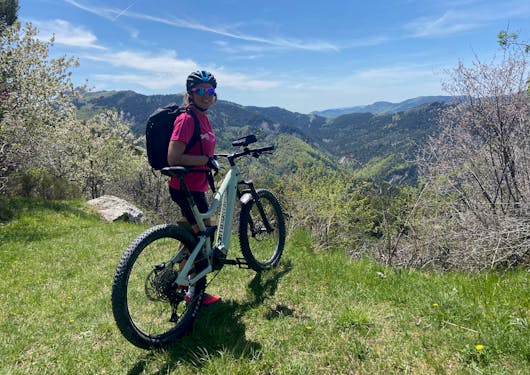 A female biker stands next to her electronic mountain bike in the French alps, smiling at the camera with mountains in the background. This is in the French Alps on a sunny day on a sustainable cycling tour.